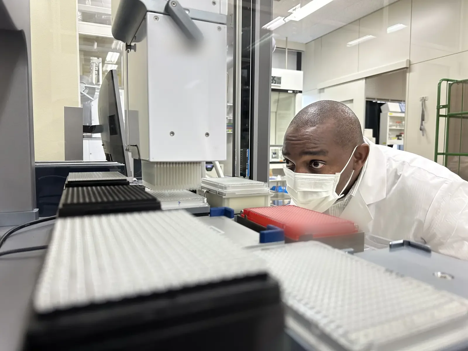A researcher in a lab coat and mask examining automated liquid-handling equipment and microplates on a laboratory bench.
